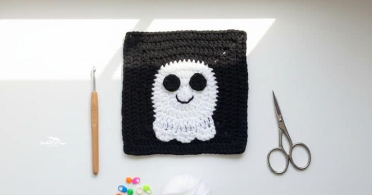 Overhead view of a black crochet granny square with a white ghost applique, styled on a white desk with a crochet hook, scissors, white yarn, and colorful stitch markers.