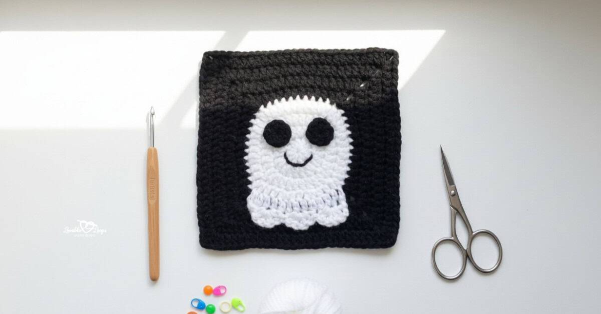 Overhead view of a black crochet granny square with a white ghost applique, styled on a white desk with a crochet hook, scissors, white yarn, and colorful stitch markers.