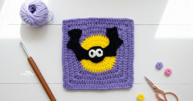 Overhead view of a purple crochet granny square with a black bat applique over a yellow moon, styled on a white wood surface with a crochet hook, purple yarn, scissors, and small crochet pieces.