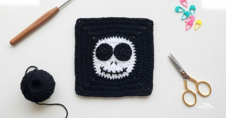 Overhead view of a black crochet granny square with a white skull face design, styled on a white desk with a crochet hook, black yarn, gold scissors, and colorful stitch markers.