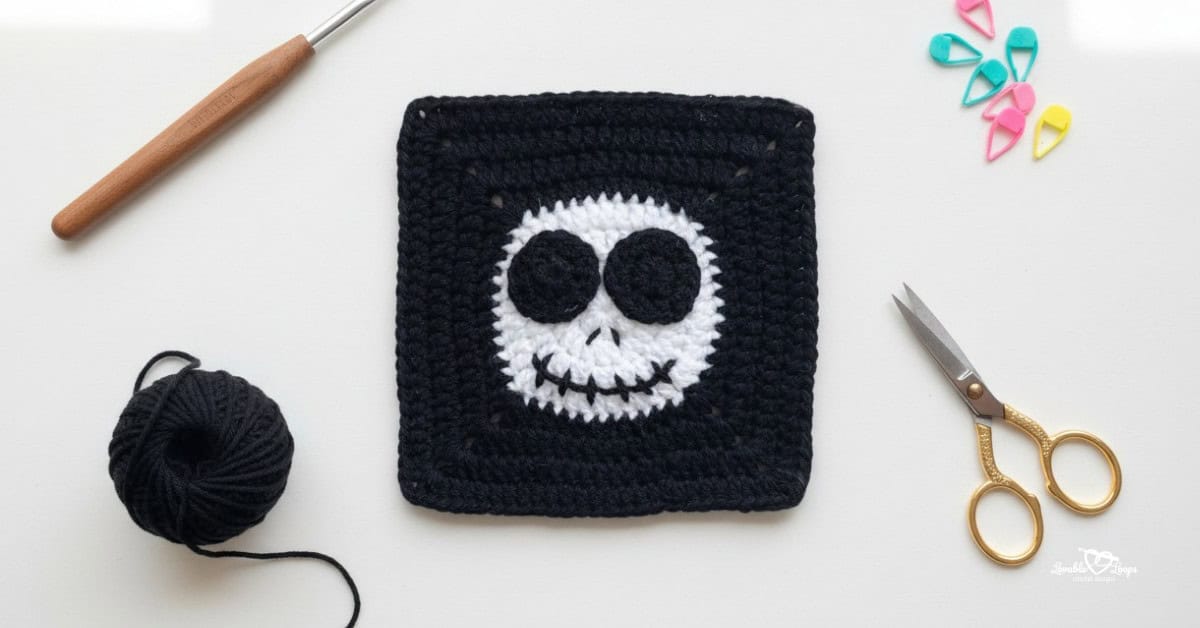 Overhead view of a black crochet granny square with a white skull face design, styled on a white desk with a crochet hook, black yarn, gold scissors, and colorful stitch markers.