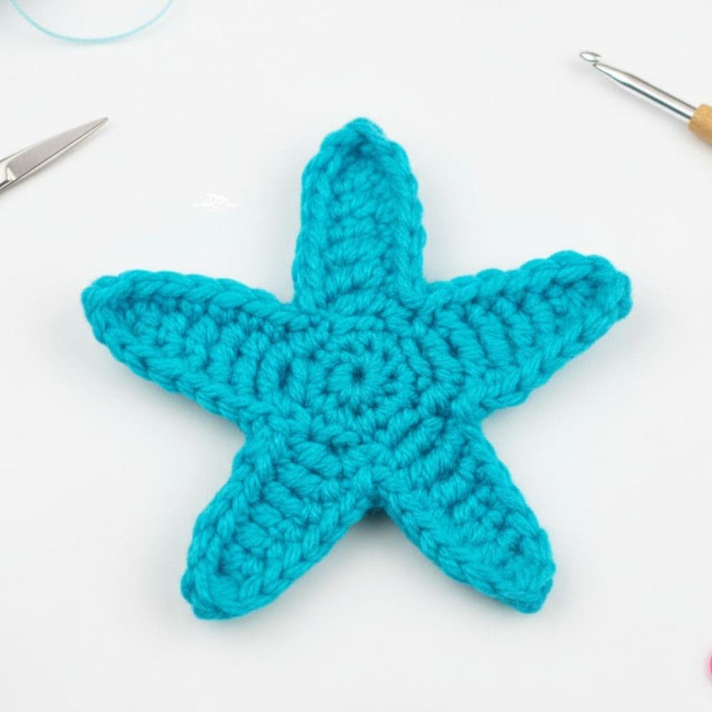 Close-up of a turquoise crochet starfish applique on a bright white work surface with simple craft tools around the edges.