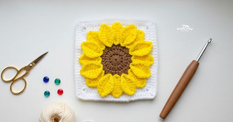 Overhead view of a white crochet granny square with a textured yellow sunflower and brown center on a white desk, styled with gold scissors, a crochet hook, yarn, and colorful gems.