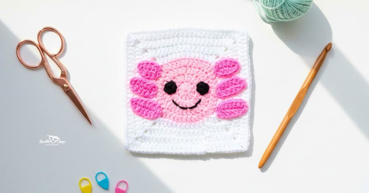 Overhead view of a white crochet square with a pink axolotl face and smile, styled on a white desk with rose gold scissors, a crochet hook, yarn, and colorful stitch markers.