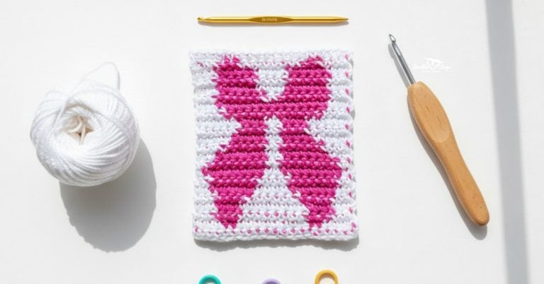 Overhead view of a white crochet square with a pink bow design, styled on a white desk with white yarn, two crochet hooks, and colorful stitch markers.