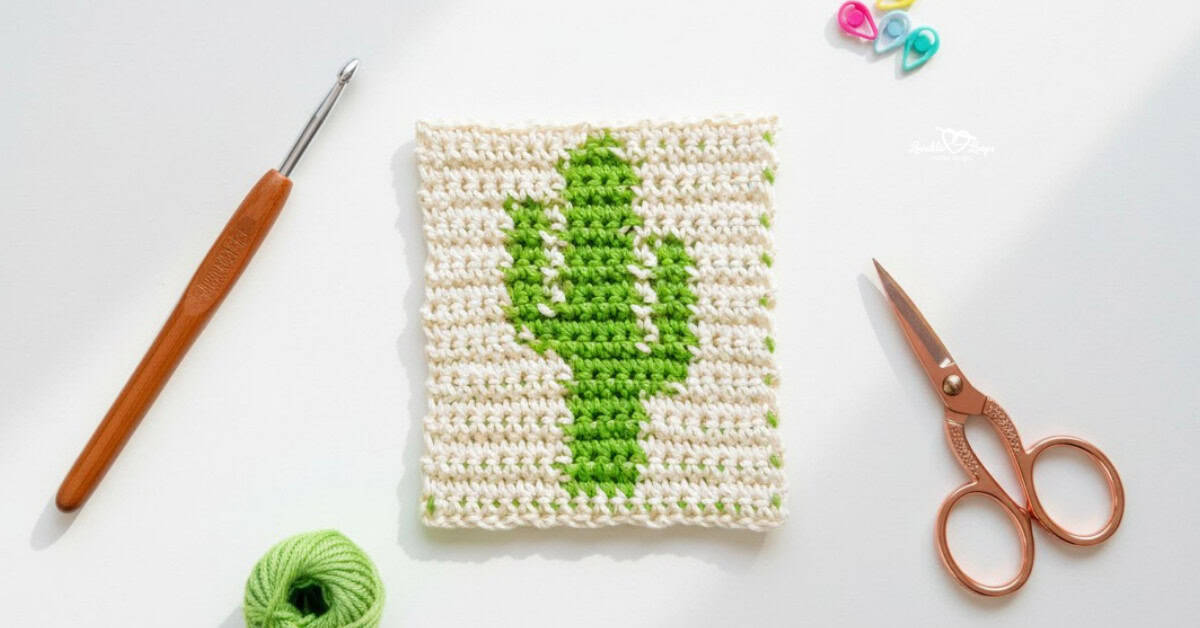 Overhead view of a cream crochet square with a green cactus design, styled on a white desk with a crochet hook, rose gold scissors, green yarn, and colorful stitch markers.