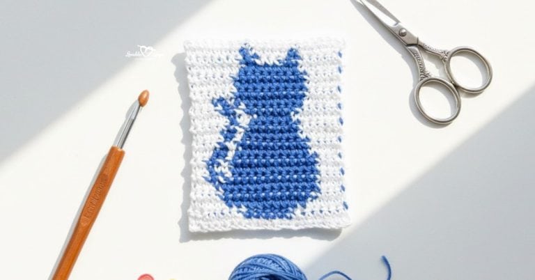 Overhead view of a white crochet square with a blue cat silhouette, styled on a white desk with a crochet hook, silver scissors, blue yarn, and colorful stitch markers.
