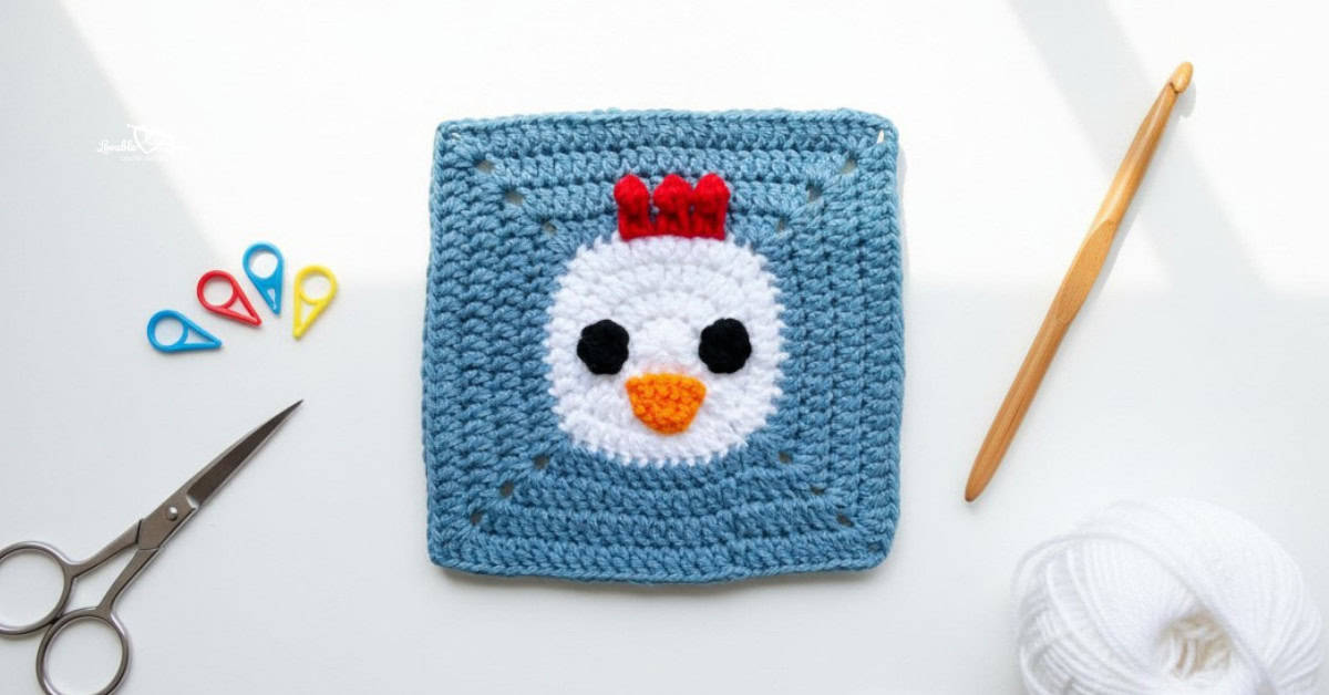 Overhead view of a blue crochet square with a chicken face applique, styled on a white desk with a crochet hook, scissors, white yarn, and colorful stitch markers.