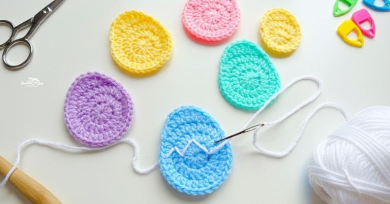 Colorful crochet egg appliques in pastel purple, yellow, pink, mint, and blue arranged on a white desktop with scissors, yarn, a crochet hook, stitch markers, and a yarn needle.