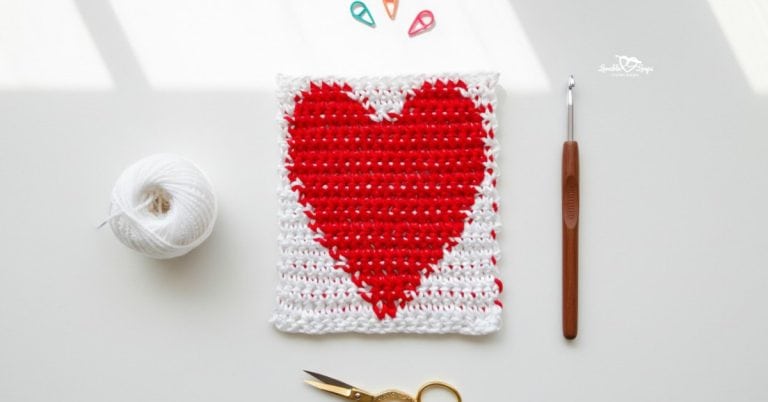 Overhead view of a white crochet square with a red heart design, styled on a white desk with white yarn, a crochet hook, gold scissors, and colorful stitch markers.