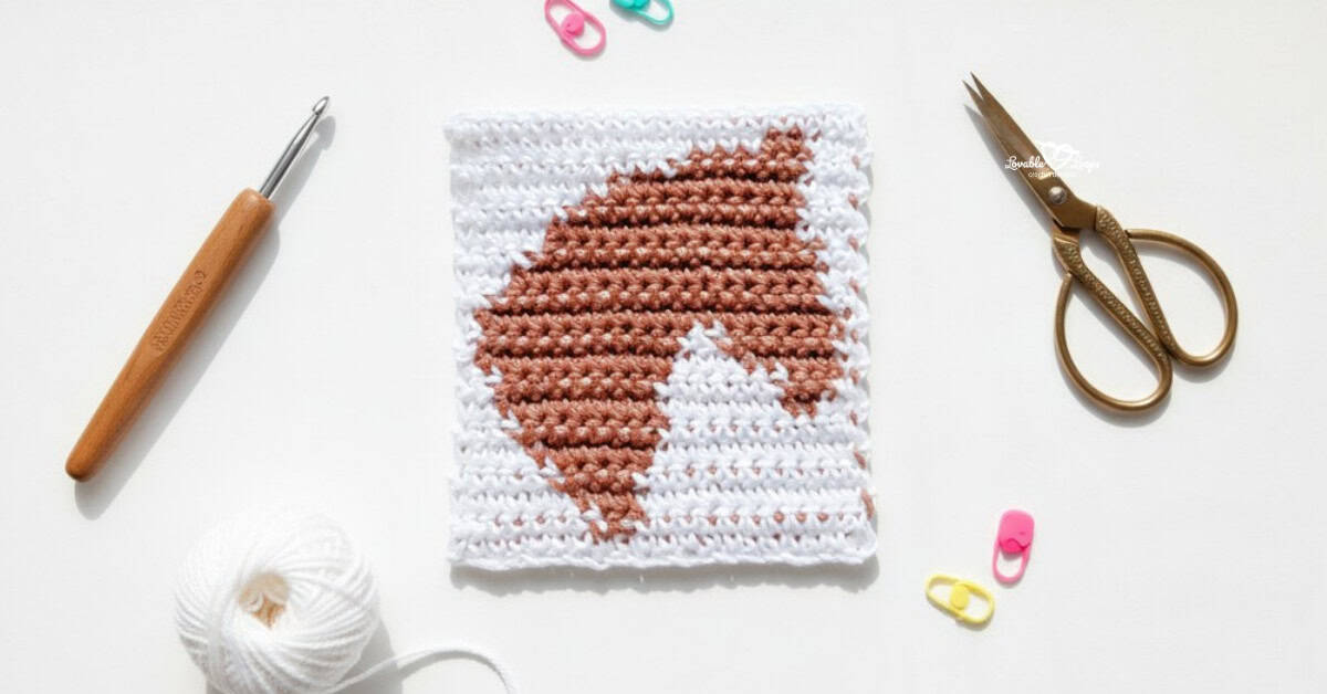 Overhead view of a white crochet square with a brown horse head design, styled on a white desk with a crochet hook, gold scissors, white yarn, and colorful stitch markers.