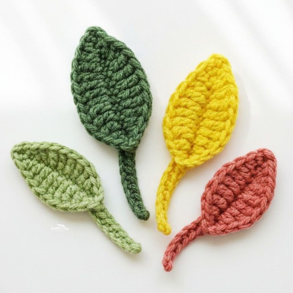 Close-up of four crochet leaf appliques in light green, dark green, yellow, and rust on a white background.
