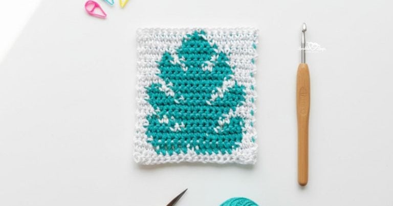 Overhead view of a white crochet square with a teal monstera leaf design, styled on a white desk with a crochet hook, teal yarn, scissors, and colorful stitch markers.