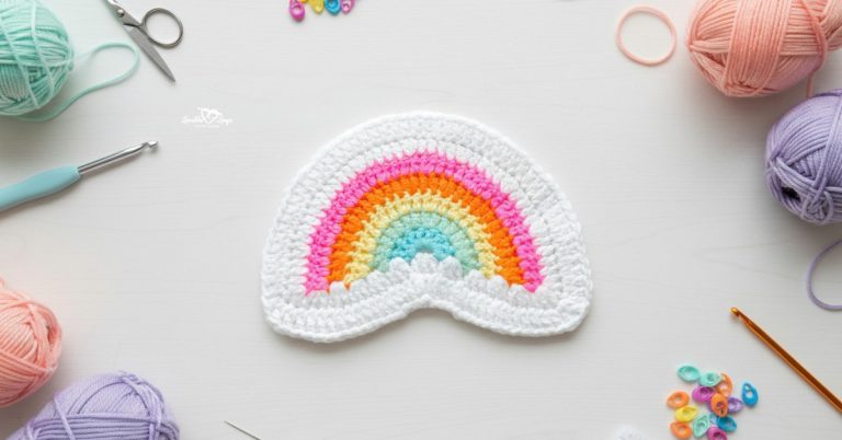 Crochet rainbow applique in pastel colors centered on a white desktop with yarn balls, crochet hooks, scissors, and colorful stitch markers around it.