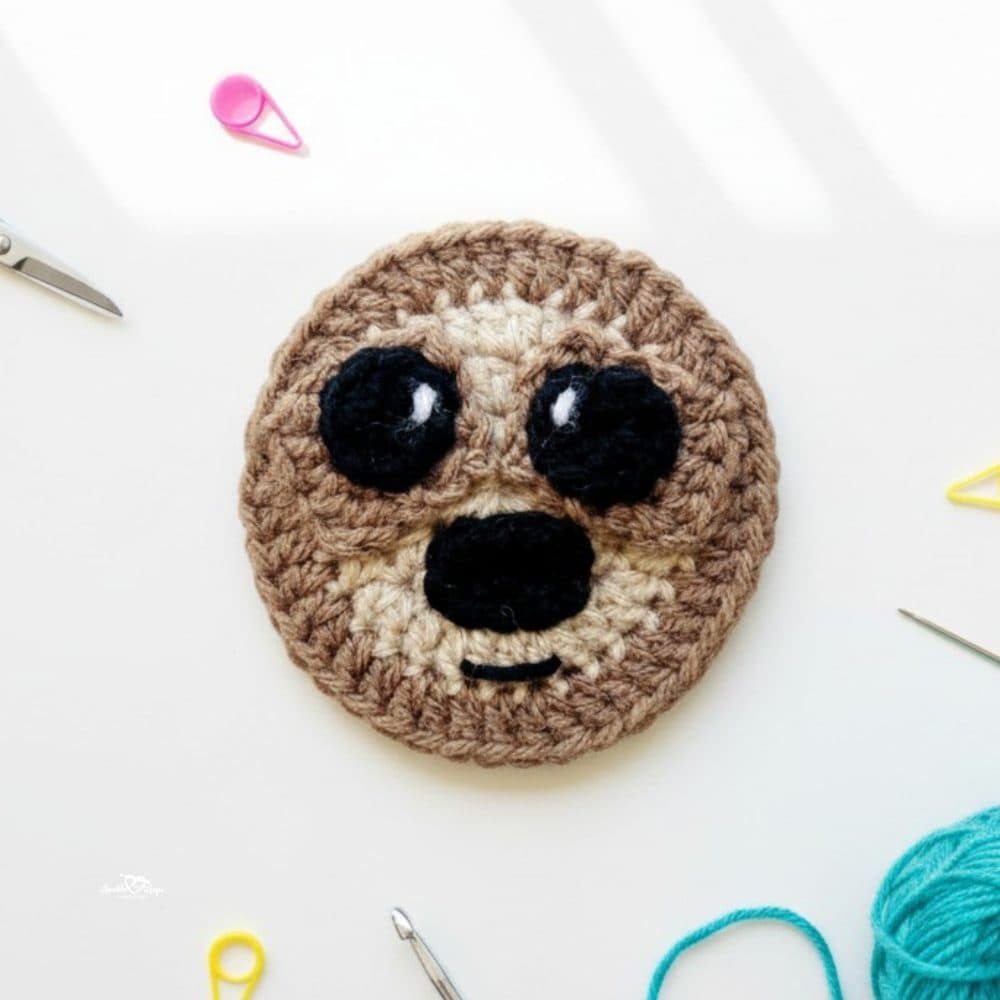 Crochet sloth applique in brown and cream yarn on a white desktop with scissors, a crochet hook, yarn, and colorful stitch markers nearby.