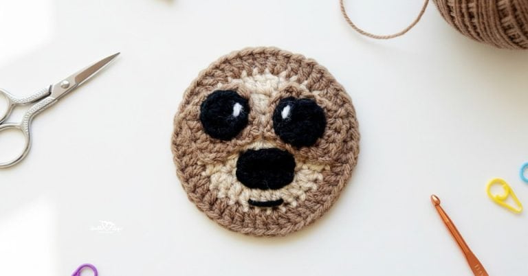 Brown crochet sloth face applique centered on a white desktop with scissors, yarn, a crochet hook, and colorful stitch markers around it.