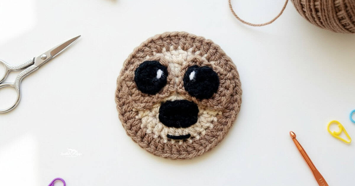 Brown crochet sloth face applique centered on a white desktop with scissors, yarn, a crochet hook, and colorful stitch markers around it.