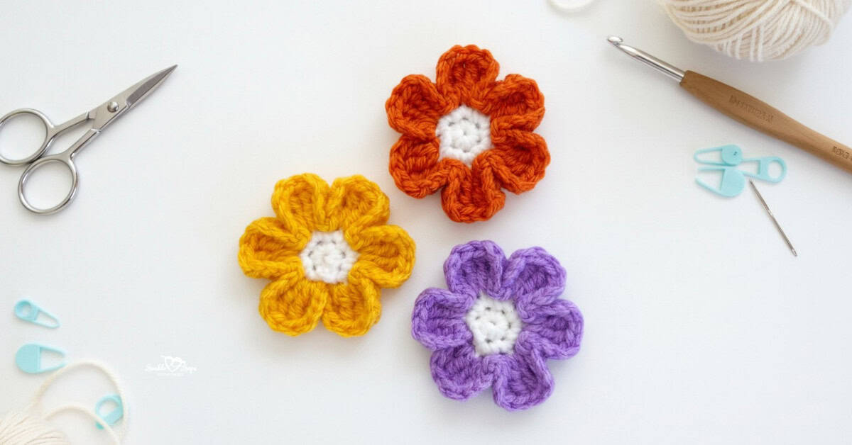 Three crochet flower appliques in yellow, orange, and purple centered on a white desktop with scissors, a crochet hook, yarn, a yarn needle, and stitch markers around the edges.