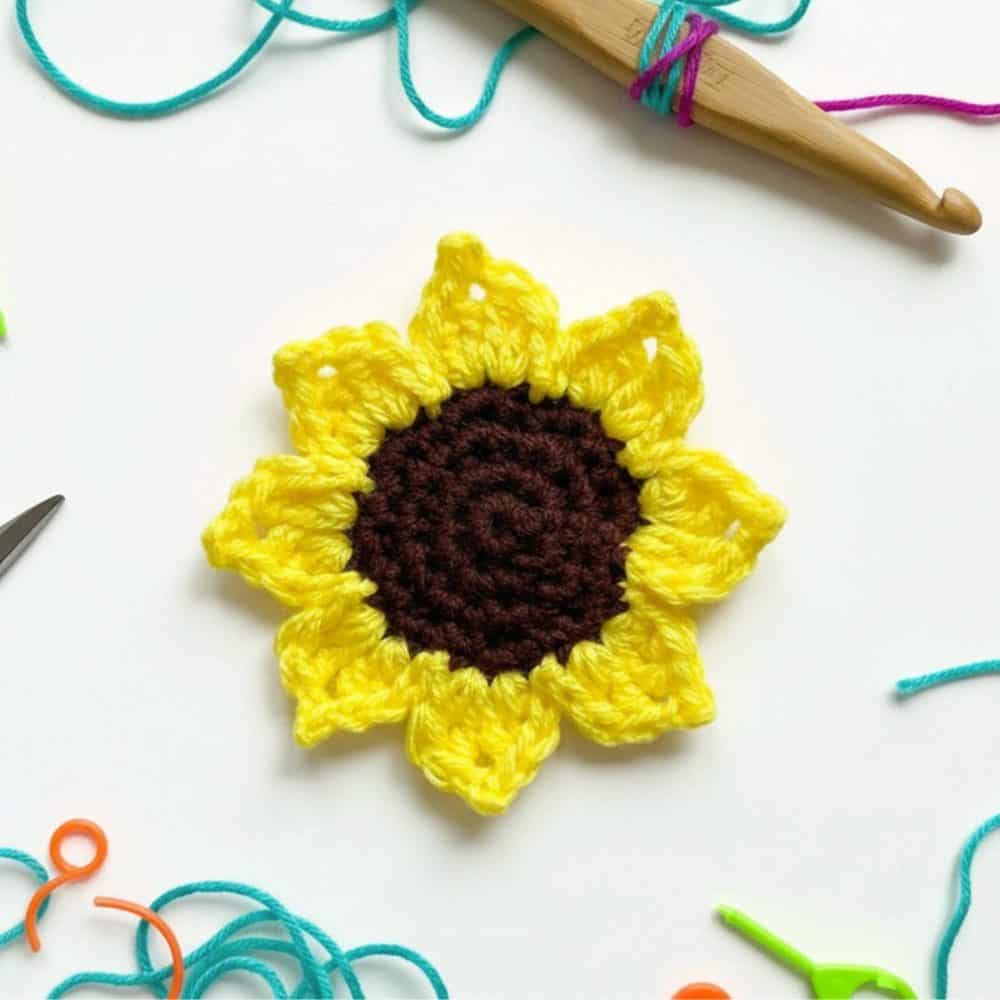 Bright yellow crochet sunflower applique with a dark brown center displayed on a white background with colorful yarn and tools around it.