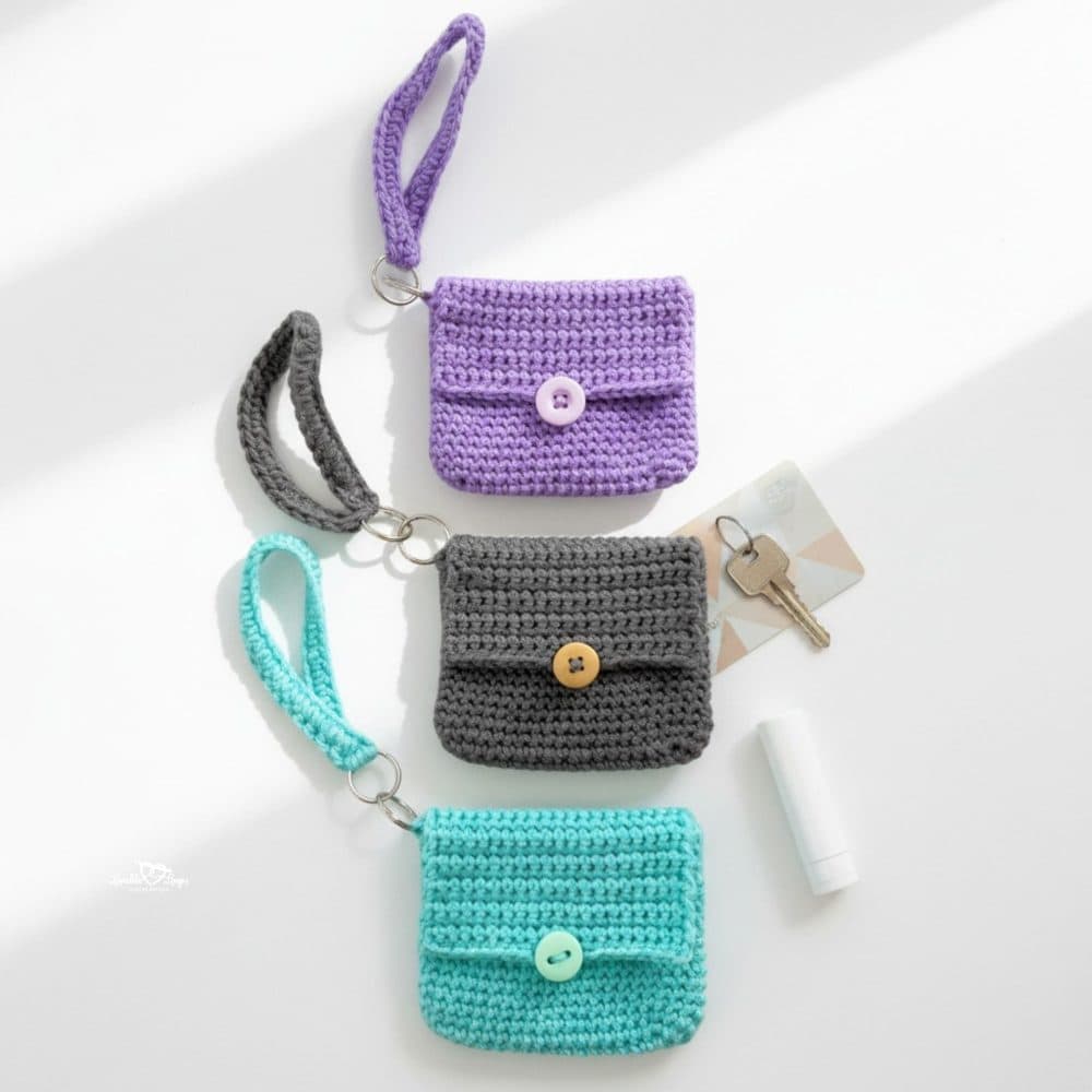 Three handmade crochet wristlet pouches in purple, gray, and turquoise with keychain rings, keys, a card, and lip balm on a white background.
