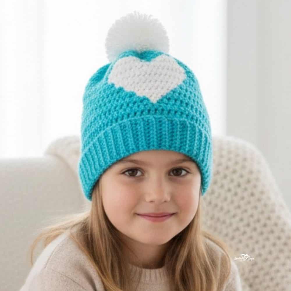 Child wearing a turquoise crochet beanie with a white heart design and a white pom pom.