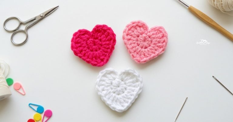 Three crochet heart appliques in pink and white centered on a white desktop with scissors, a crochet hook, colorful stitch markers, and yarn needles around the edges.