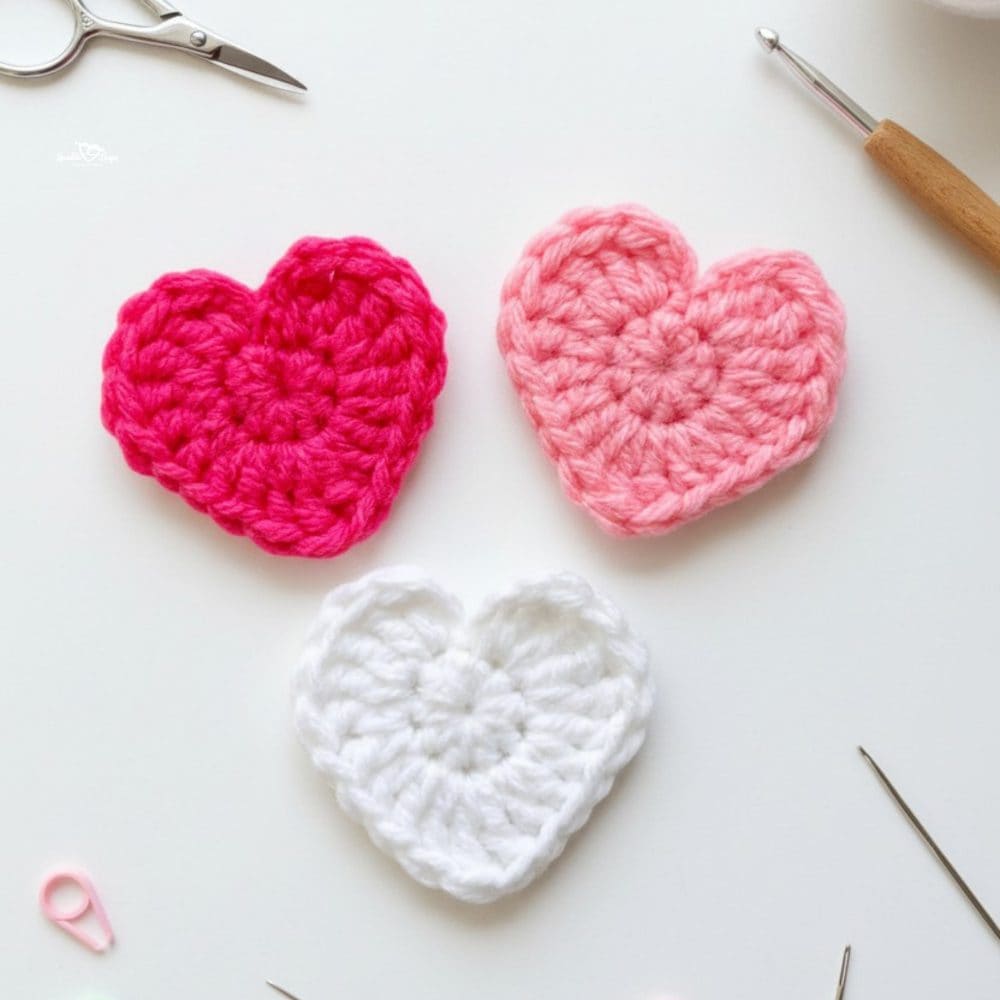 Three small crochet heart appliques in hot pink, light pink, and white yarn on a white desktop with scissors, a crochet hook, yarn, and yarn needles nearby.