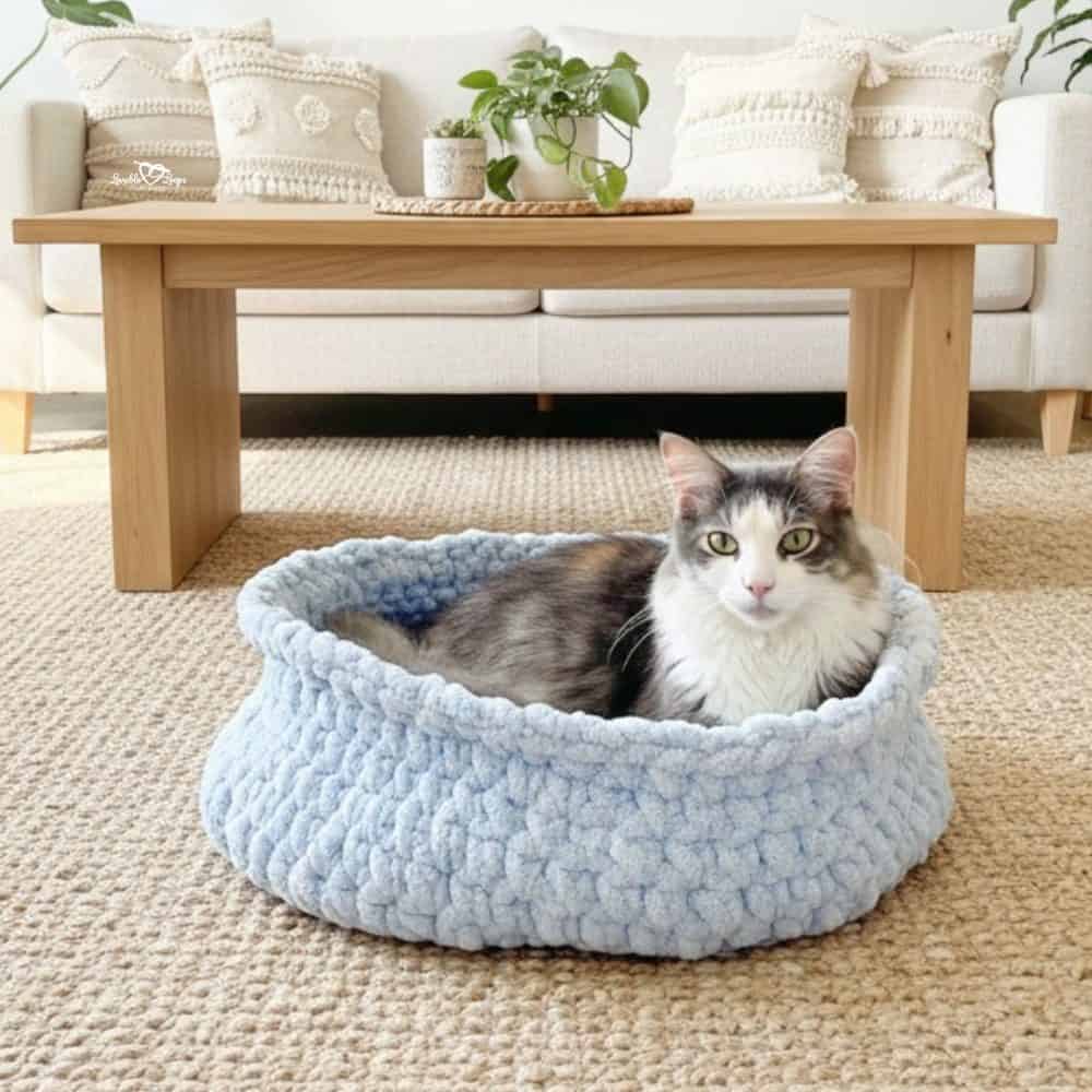 Gray and white cat lying in a soft blue crochet cat bed made with plush yarn in a cozy neutral living room.