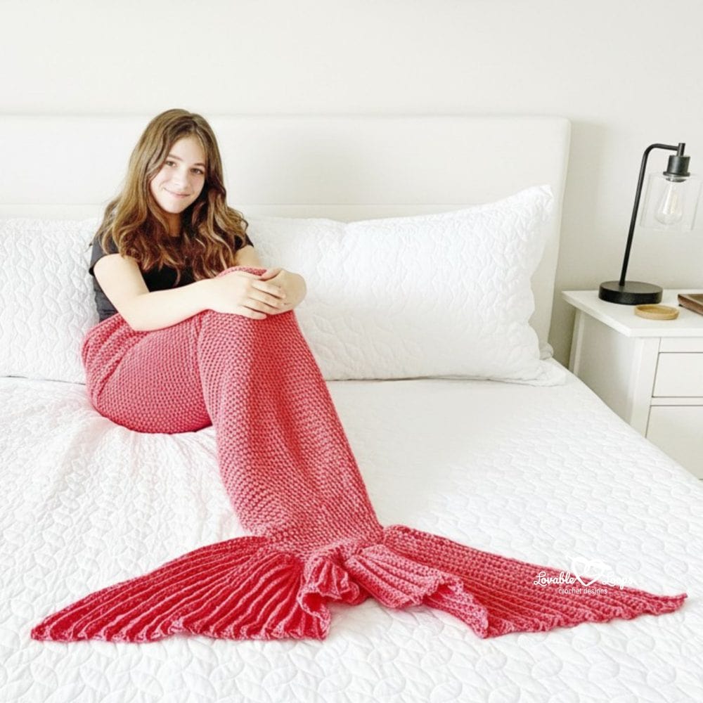 Girl sitting on a white bed wrapped in a pink crochet mermaid tail blanket with a ribbed textured fin.