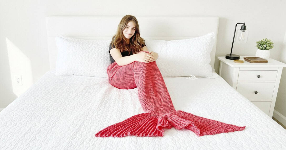 Girl sitting on a white bed wrapped in a pink crochet mermaid tail blanket with a textured fin.
