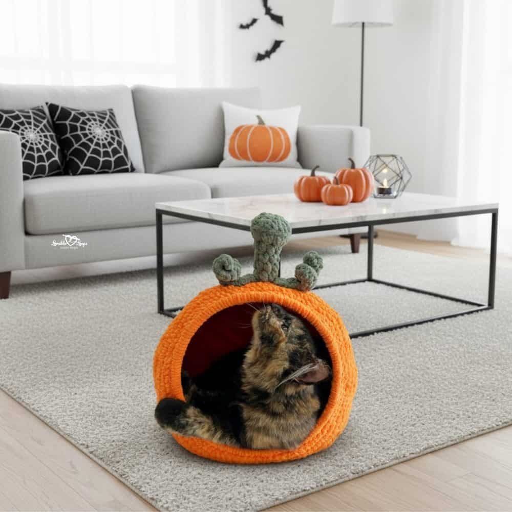 Tortoiseshell cat sitting inside an orange pumpkin crochet cat pod in a bright living room with Halloween decor.