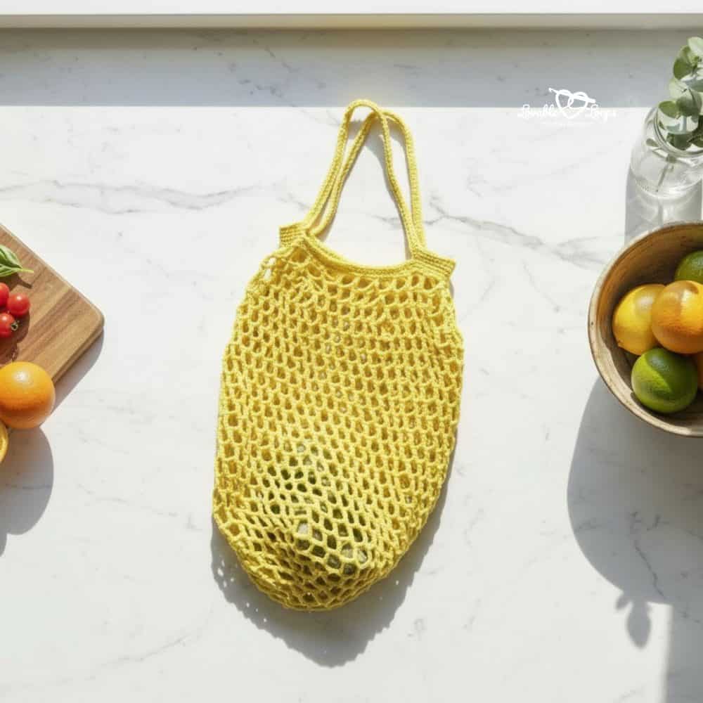 Yellow mesh crochet produce bag lying on a white marble counter with citrus fruit, tomatoes, basil, and a bowl of lemons and limes nearby.