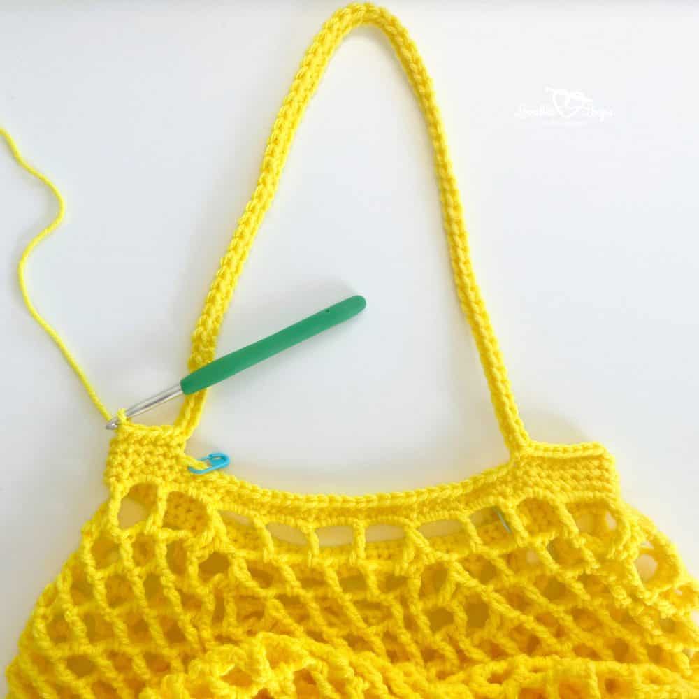 Yellow crochet produce bag in progress with a green crochet hook, stitch marker, and long strap on a white background.