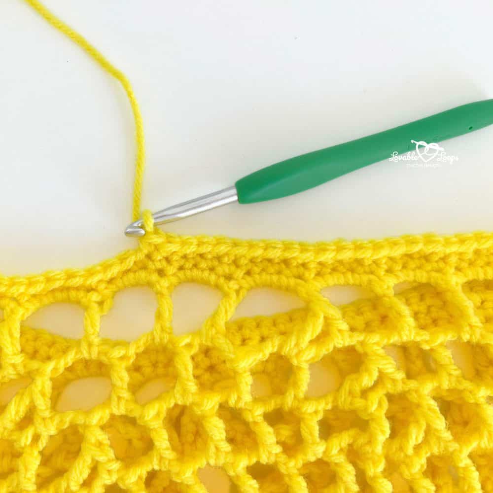Close-up of a yellow crochet produce bag being worked with a green crochet hook along the top edge.