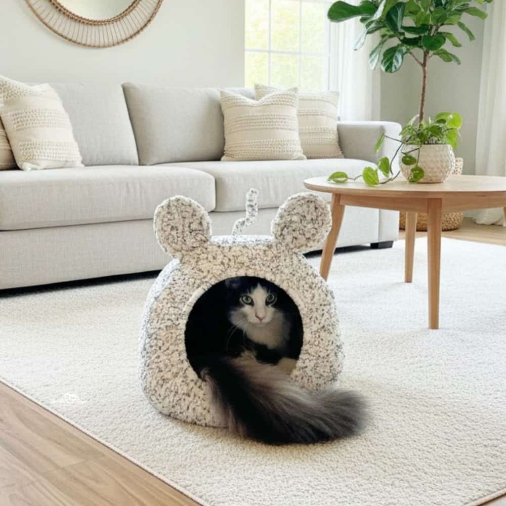 Black and white cat sitting inside a cream speckled mouse ears crochet cat pod in a bright neutral living room.