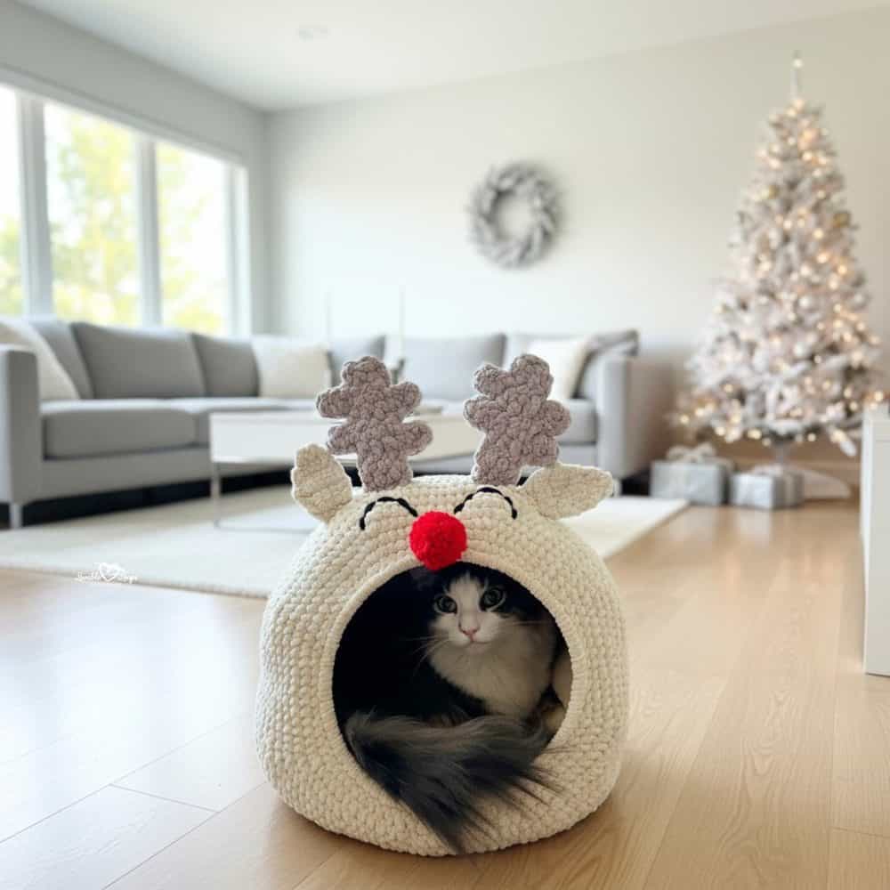 Cream reindeer crochet cat pod with antlers and a red nose, styled in a bright holiday living room with a cat peeking out.