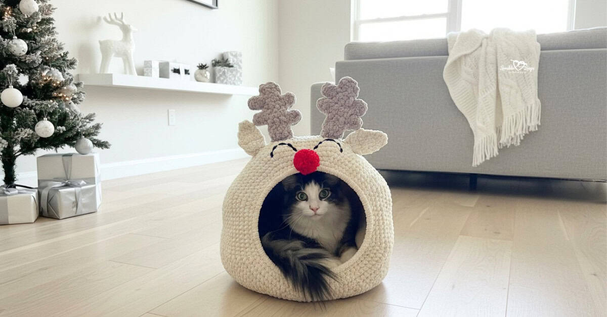 Black and white cat sitting inside a cream reindeer crochet cat pod with antlers, ears, closed eyes, and a red pom-pom nose in a bright Christmas living room.