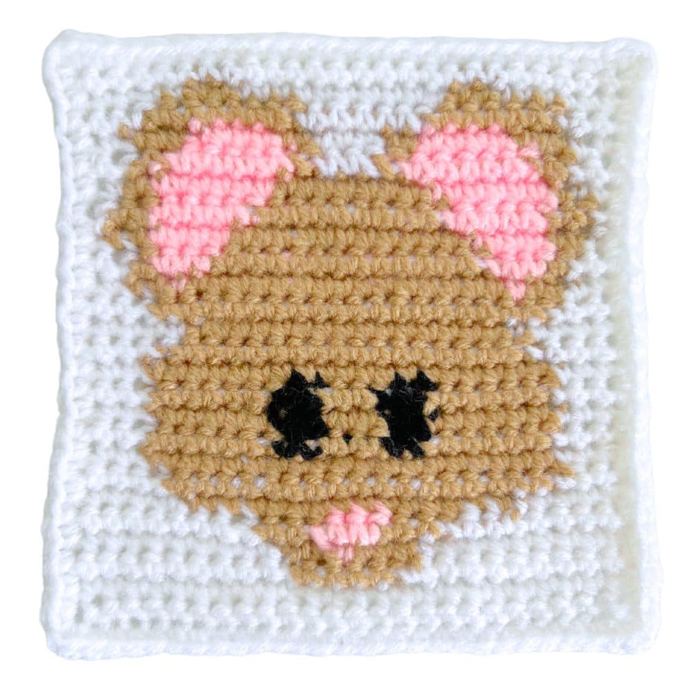 Close-up photo of a handmade crochet mouse square with a tan mouse face, pink inner ears, black eyes, and a pink nose on a white crocheted background.