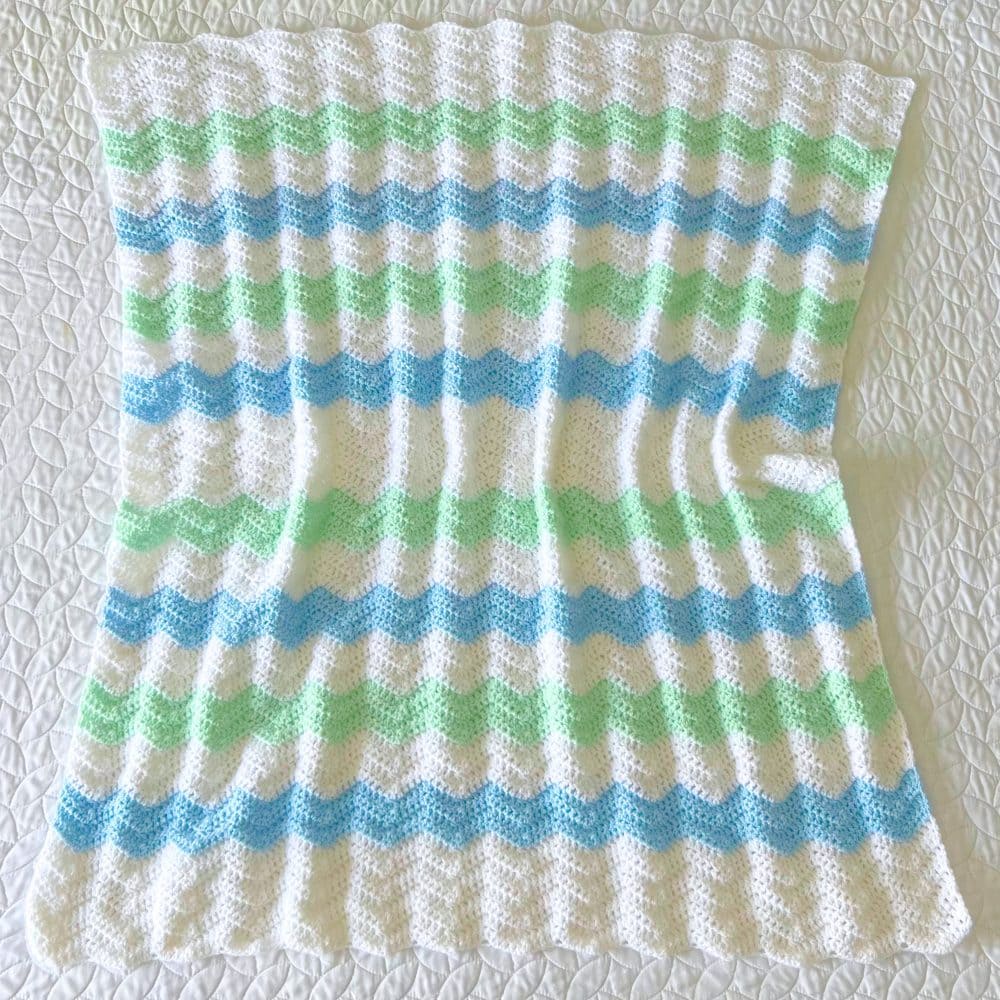 Crochet chevron baby blanket with white, blue, and green ripple stripes laid flat on a white quilt.