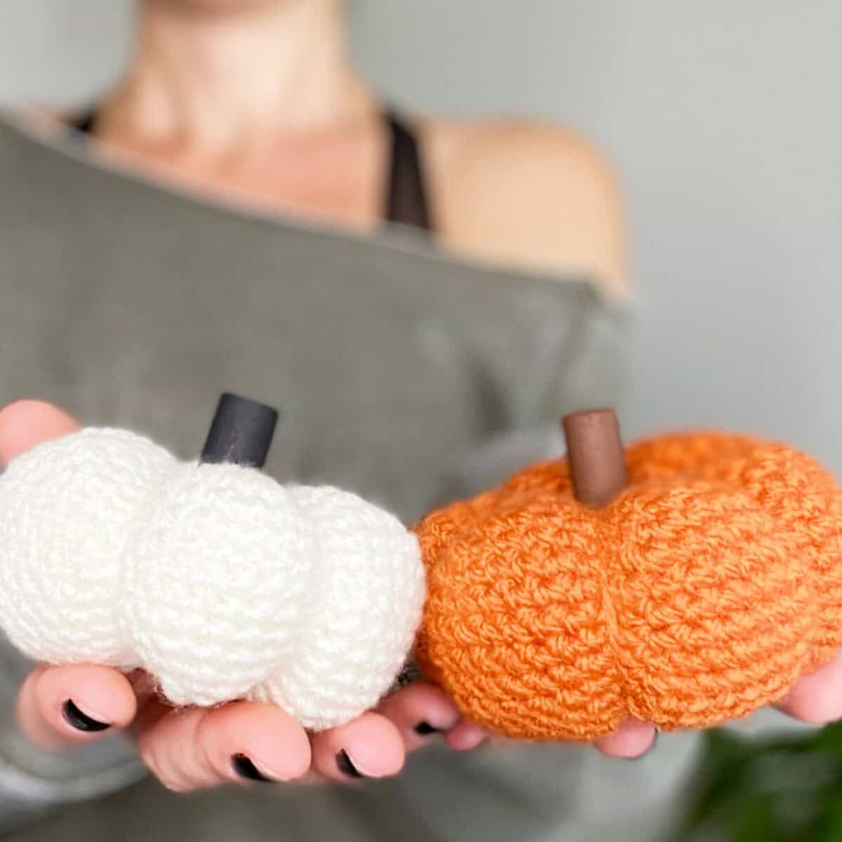 A person holding two handmade crochet pumpkins, one white and one orange, with stems, against a soft blurred background.