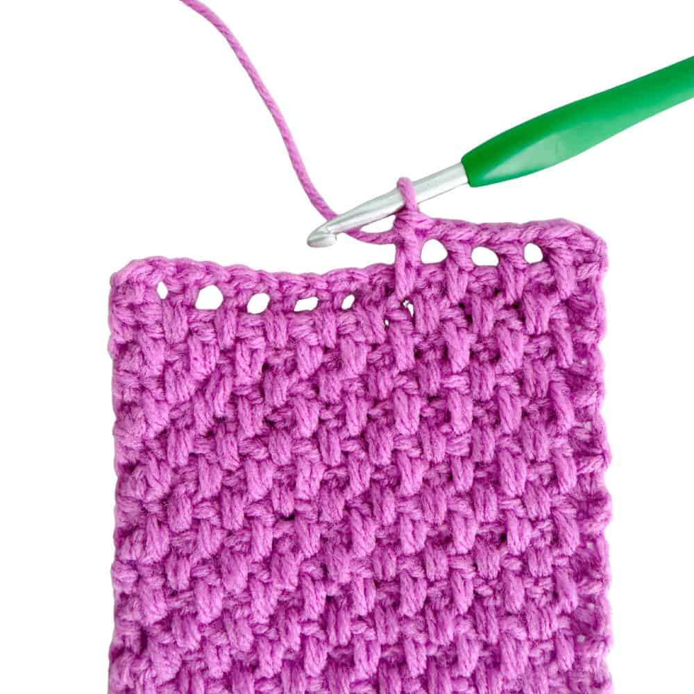 Close-up of a pink crochet moss stitch scarf in progress, showing the textured stitch pattern and a green-handled crochet hook.