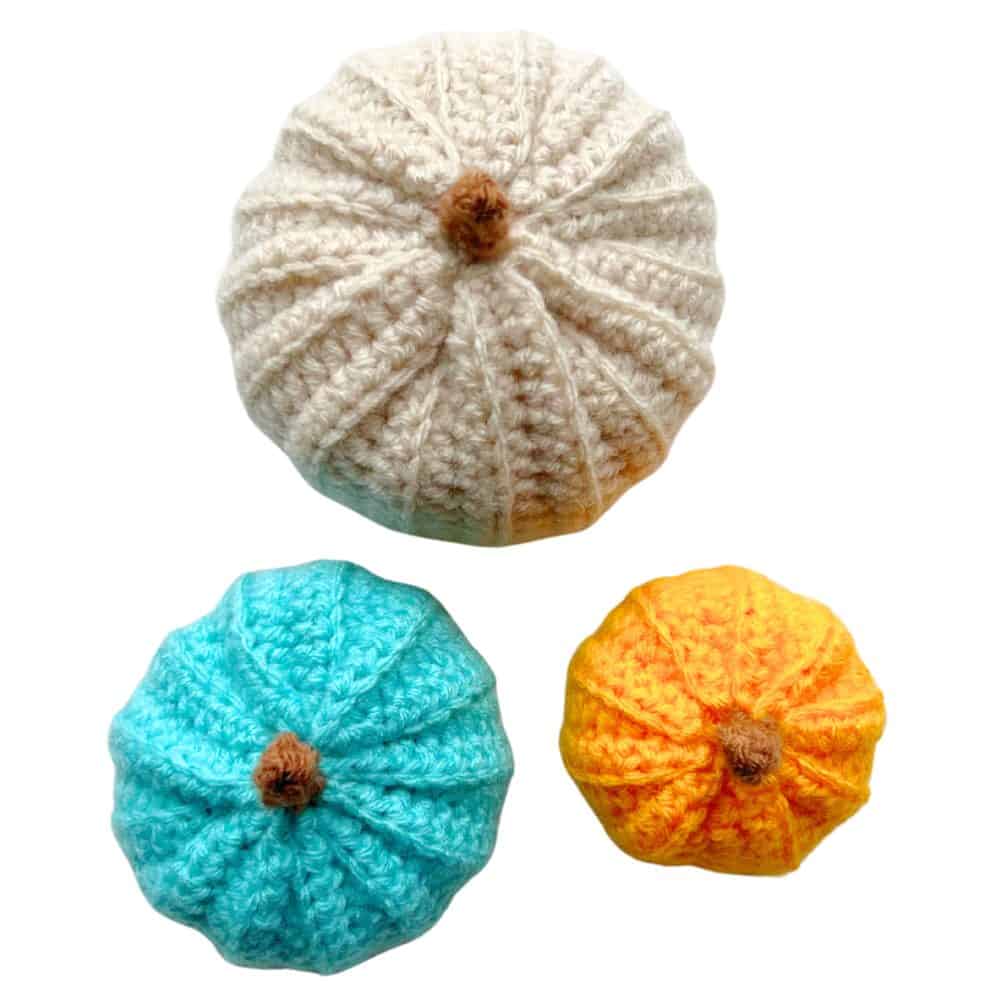 Three finished crochet pumpkins in cream, turquoise, and orange with ribbed texture and small brown stems on a white background.