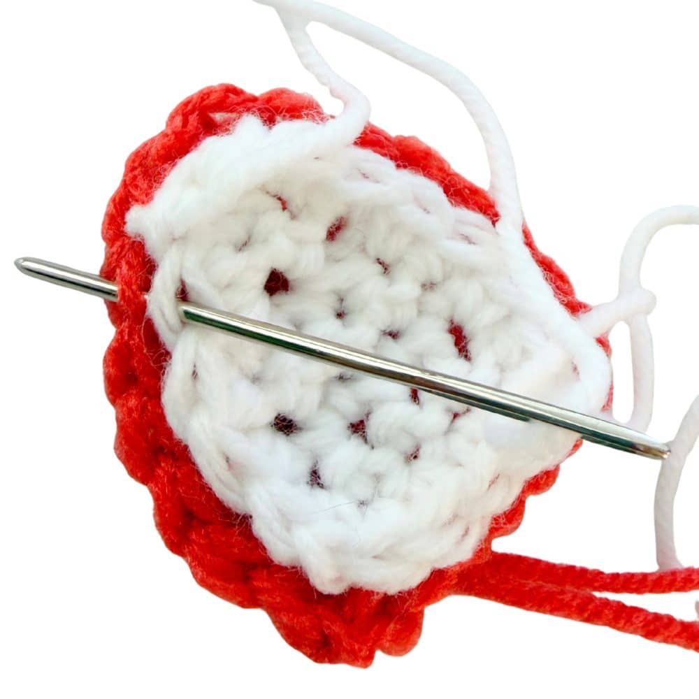 Close-up showing a white crochet inner fox ear being sewn to an orange outer ear with a yarn needle through the edge stitches.